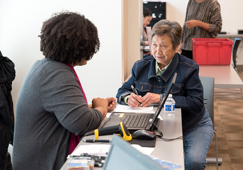 Two adults seated at a computer, one with pen in hand.