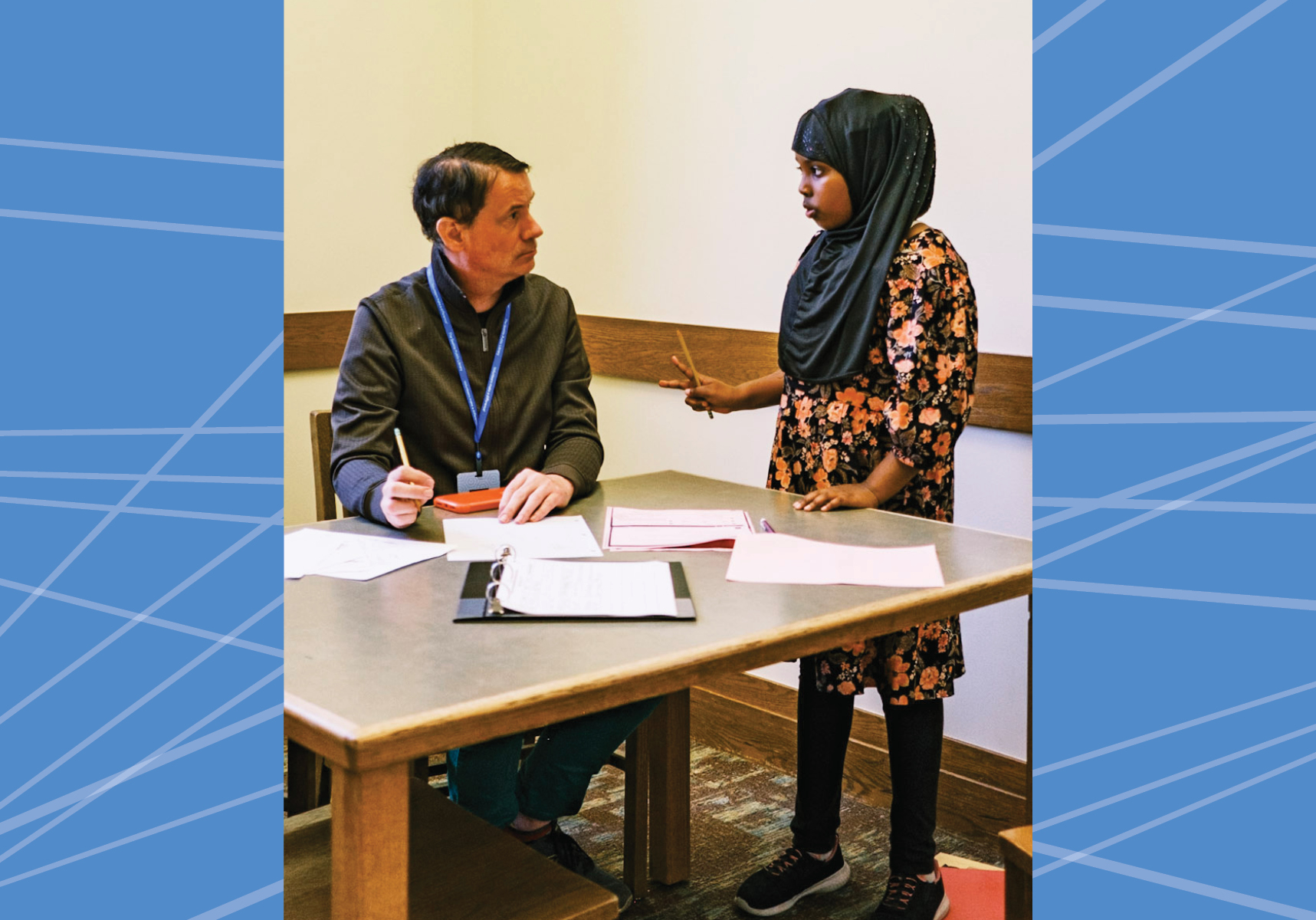 An adult sitting at a table talking to a youth standing next to the table.